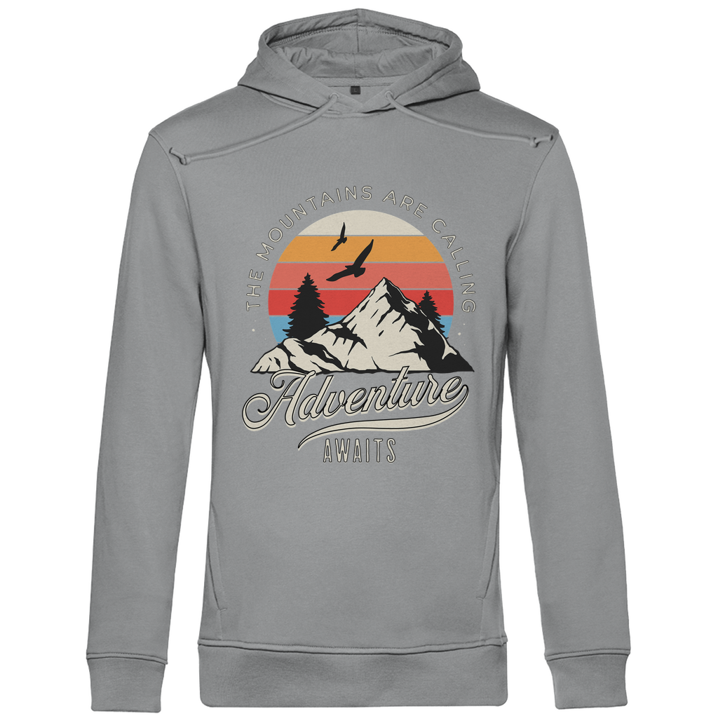 Ein hellgrauer Hoodie mit einem Outdoor-Design, das einen Berg, Bäume und fliegende Vögel zeigt. Im Hintergrund ist ein Sonnenuntergang in warmen Farbstreifen abgebildet. Der Text "The Mountains Are Calling" ist im oberen Bereich des Designs zu sehen, während "Adventure Awaits" in geschwungener Schrift darunter steht. Das Design vermittelt eine abenteuerliche und naturverbundene Atmosphäre, ideal für Liebhaber von Outdoor-Aktivitäten und Bergen.