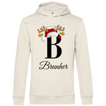 Ein cremefarbender Hoodie mit einem weihnachtlichen Design. Im Zentrum steht ein großer schwarzer Buchstabe "B", der oben mit einem Weihnachtsmann-Hut und Rentiergeweihen geschmückt ist, an denen bunte Christbaumkugeln hängen. Unter dem Buchstaben steht der personalisierte Name "Brunher" in eleganter, geschwungener Schrift. Das Design bringt eine festliche Stimmung und lässt sich individuell mit einem Namen anpassen.