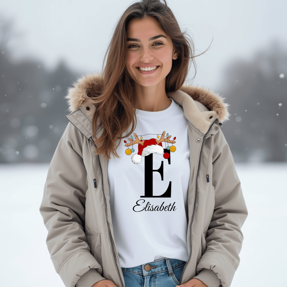 Eine Frau trägt ein weißes T-Shirt mit einem weihnachtlichen Design des Buchstabens 'E' in schwarzer Schrift. Der Buchstabe ist verziert mit einem Weihnachtsmann-Hut und Rentiergeweihen, an denen bunte Kugeln hängen. Darunter steht der Name 'Elisabeth' in eleganter, geschwungener Schrift. Sie trägt dazu eine warme, beige Winterjacke mit Kapuze und Fellbesatz und steht vor einer winterlichen Landschaft. Das T-Shirt vermittelt eine festliche und personalisierte Stimmung, perfekt für die Weihnachtszeit. 