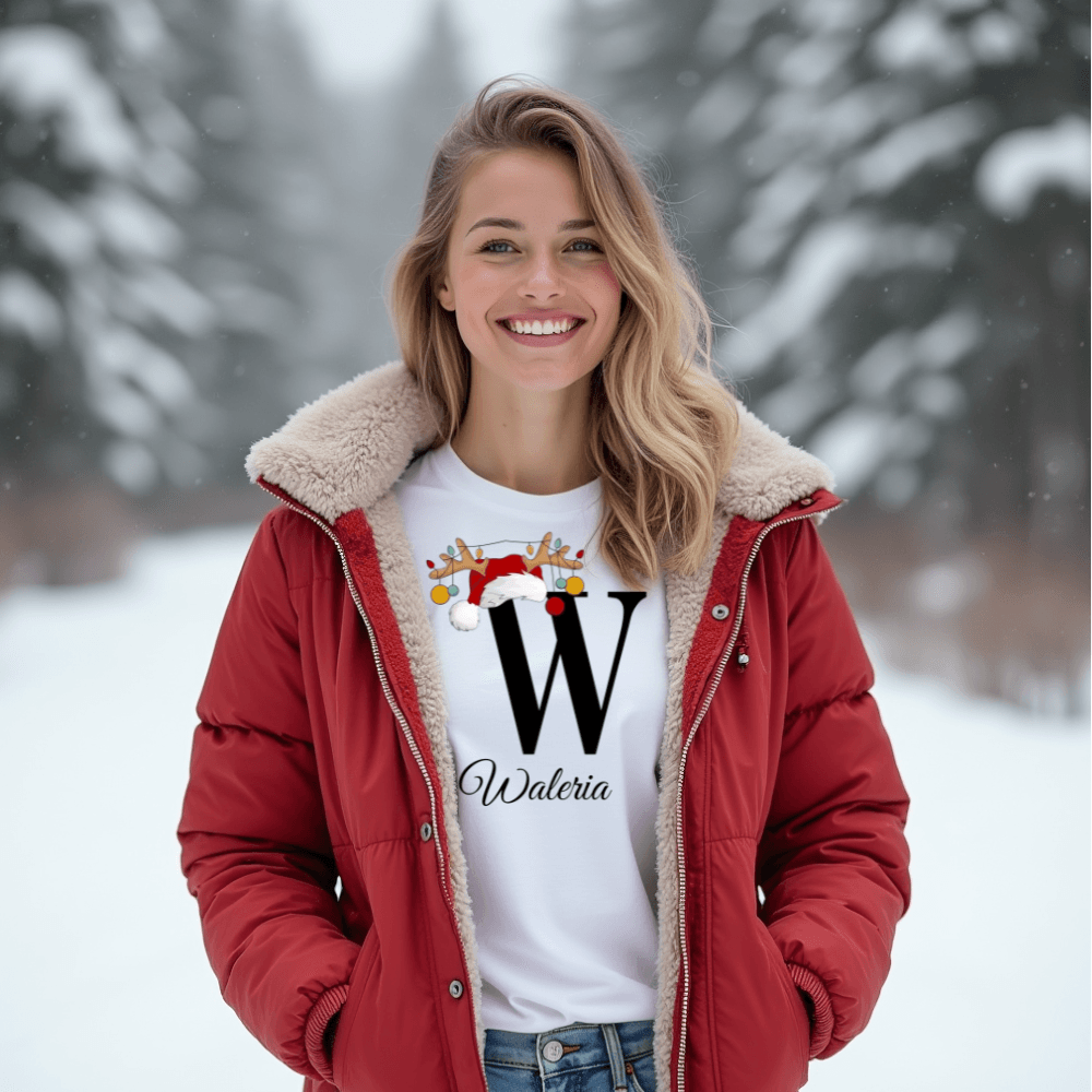 Eine Frau trägt ein weißes T-Shirt mit einem großen schwarzen Buchstaben 'W', verziert mit einem Weihnachtsmann-Hut und Rentiergeweih, an dem bunte Kugeln hängen. Unter dem Buchstaben steht der Name 'Waleria' in eleganter Schrift. Die winterliche Szenerie im Hintergrund unterstreicht die festliche, weihnachtliche Stimmung des Designs.