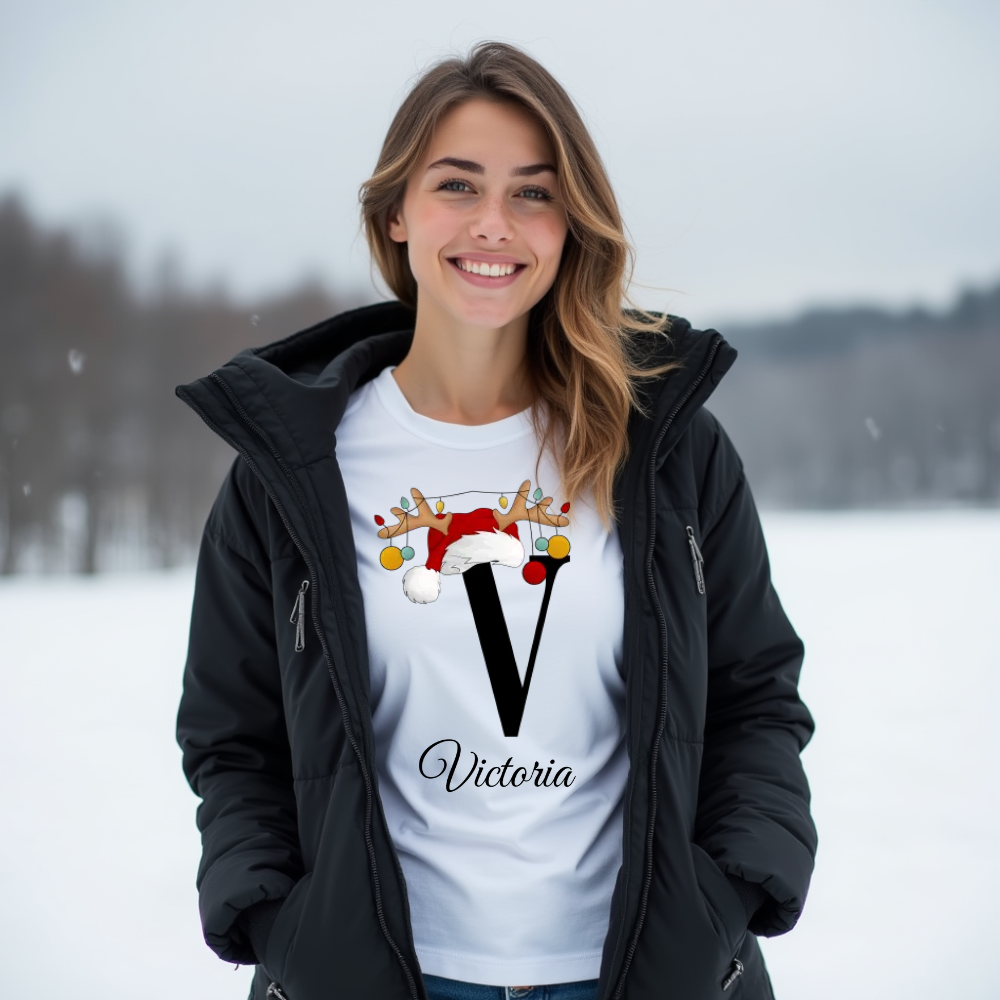 Weißes T-Shirt mit einem großen schwarzen Buchstaben 'V', verziert mit einem Weihnachtsmann-Hut und Rentiergeweihen, an denen bunte Kugeln hängen. Darunter ist der Name 'Victoria' in eleganter, geschwungener Schrift zu sehen. Das Design strahlt eine festliche und persönliche Note aus.