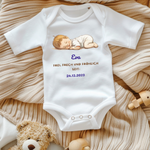 Ein weißer Baby-Body mit einem niedlichen Motiv eines schlafenden Babys. Darüber ist der Name 'Eva' in blauer Schrift zu sehen. Darunter steht der Text 'Frei, frech und fröhlich seit: 24.12.2023' in goldener und blauer Schrift. Der Body liegt auf einer beigen, gestrickten Decke und wird von Spielzeug umgeben, was eine warme und gemütliche Stimmung vermittelt. Ein perfektes, personalisiertes Geschenk zur Geburt.