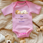 Ein rosa Baby-Body mit einem niedlichen Motiv eines schlafenden Babys. Darüber ist der Name 'Eva' in blauer Schrift zu sehen, gefolgt vom Text 'Frei, frech und fröhlich seit: 24.12.2023' in goldener und blauer Schrift. Der Body liegt auf einer beigen, gestrickten Decke und wird von Spielzeug umrahmt, was eine warme und heimelige Atmosphäre vermittelt. Ein liebevoll personalisiertes Geschenk zur Geburt.