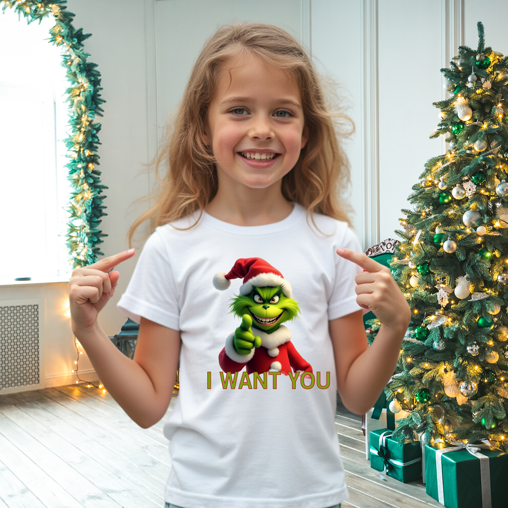 Ein Mädchen trägt ein weißes T-Shirt mit einem lustigen, weihnachtlichen Motiv. Auf dem T-Shirt ist eine grüne Figur mit Weihnachtsmann-Mütze abgebildet, die in die Kamera zeigt, darunter steht der Text "I WANT YOU". Das Mädchen lächelt und zeigt auf das Motiv. Im Hintergrund steht ein geschmückter Weihnachtsbaum, der eine festliche Atmosphäre schafft.