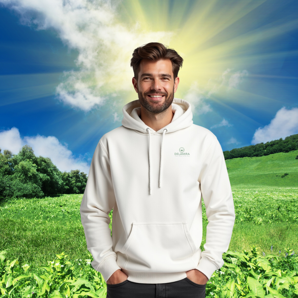 Ein Mann trägt einen weißen Premium Bio Hoodie mit delamira-Logo, stehend auf einer grünen Wiese unter einem sonnigen Himmel. Nachhaltige Mode in natürlicher Umgebung.