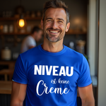 Ein Mann in einem blauen T-Shirt mit dem humorvollen Aufdruck 'Niveau ist keine Creme', lächelnd in einem gemütlichen Café-Ambiente.