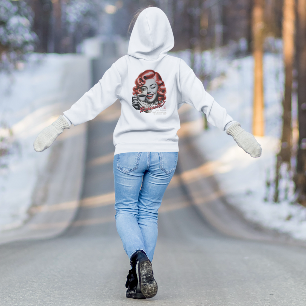 Eine Person trägt einen weißen Hoodie mit einem Vintage-inspirierten Design auf der Rückseite. Das Motiv zeigt eine Frau mit roten Locken, die eine Kaffeetasse hält und zwinkert, begleitet vom Text „None of my Business!“. Die Person läuft auf einer verschneiten Straße, umgeben von winterlicher Landschaft, mit ausgebreiteten Armen und trägt weiße Fäustlinge, Jeans und Winterstiefel.