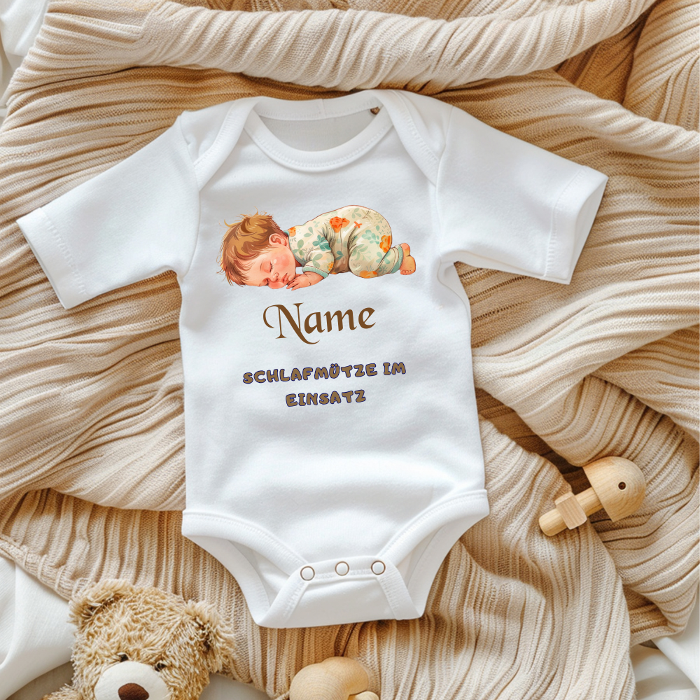 Ein weißer Baby-Body mit einem süßen Motiv eines schlafenden Babys in gemusterter Schlafkleidung. Darüber steht der personalisierbare Schriftzug 'Name' in brauner, verspielter Schrift. Darunter befindet sich der Text 'SCHLAFMÜTZE IM EINSATZ' in grauer, verspielter Schrift. Der Body liegt auf einer beigen, gestrickten Decke, umgeben von Spielzeug, was eine gemütliche und liebevolle Atmosphäre schafft. Ein perfektes, personalisierbares Geschenk für kleine Schlafmützen.