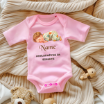 Ein rosa Baby-Body mit einem süßen Motiv eines schlafenden Babys in gemusterter Schlafkleidung. Darüber steht der personalisierbare Schriftzug 'Name' in brauner, verspielter Schrift. Darunter befindet sich der Text 'SCHLAFMÜTZE IM EINSATZ' in grauer, verspielter Schrift. Der Body liegt auf einer beigen, gestrickten Decke, umgeben von Spielzeug, was eine gemütliche und liebevolle Atmosphäre schafft. Ein perfektes, personalisierbares Geschenk für kleine Schlafmützen.