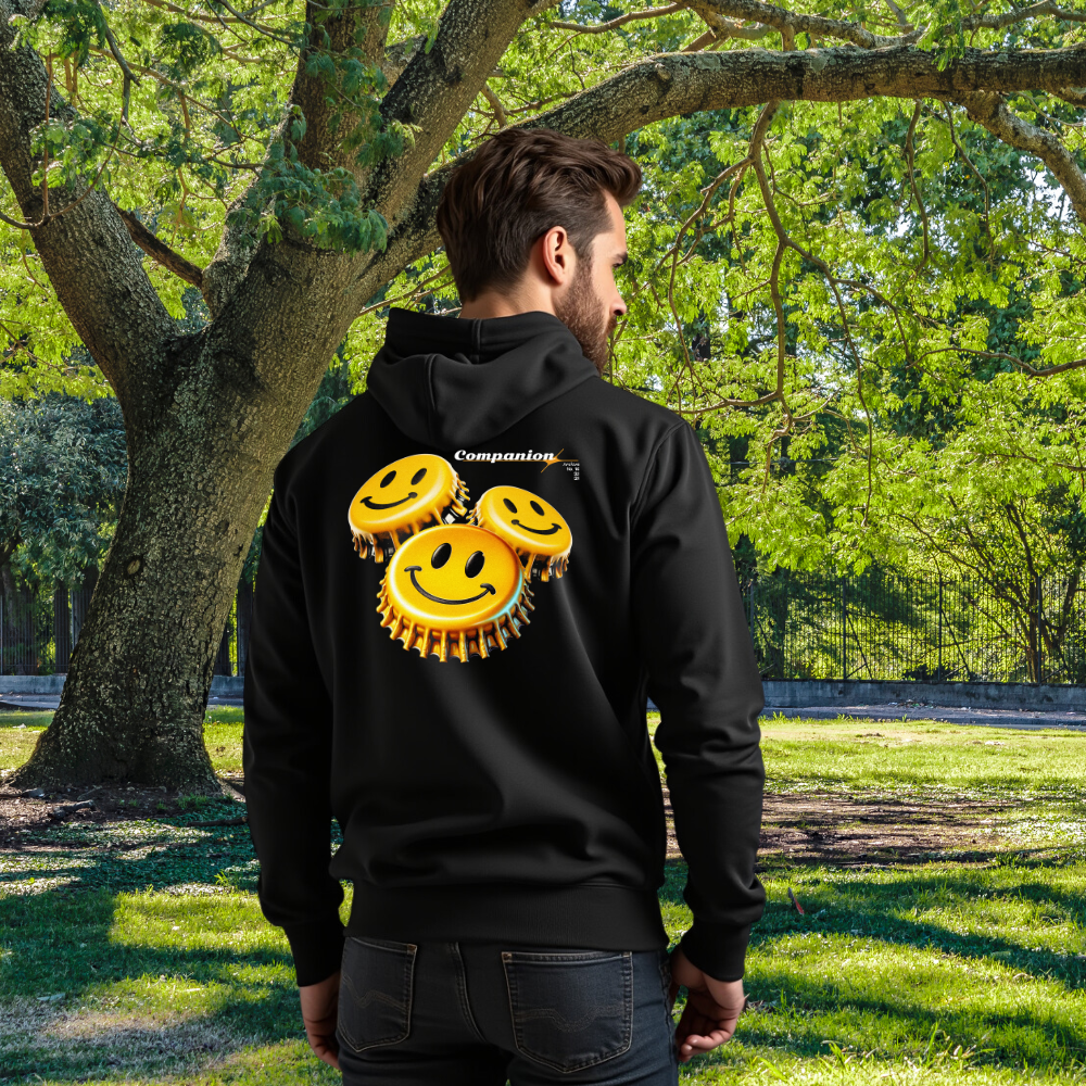 Ein Mann steht im Freien und trägt einen schwarzen Hoodie aus Bio-Baumwolle, dessen Rückseite ein auffälliges Design aus drei gelben Smiley-Gesichtern auf Kronkorken zeigt. Oben rechts ist der Schriftzug 'Companion' zu sehen. Der hochwertige Hoodie passt sich gut an und strahlt eine fröhliche und moderne Ästhetik aus.