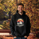 Ein Mann im schwarzen Hoodie mit einem Outdoor-Design, das einen Berg mit Bäumen und fliegenden Vögeln zeigt. Im Hintergrund ist ein Sonnenuntergang in warmen Farben abgebildet. Oberhalb des Designs steht der Text "The Mountains Are Calling" und darunter "Adventure Awaits" in geschwungener Schrift. Der herbstliche Hintergrund mit gelben und orangen Blättern verstärkt die naturverbundene und abenteuerliche Stimmung.