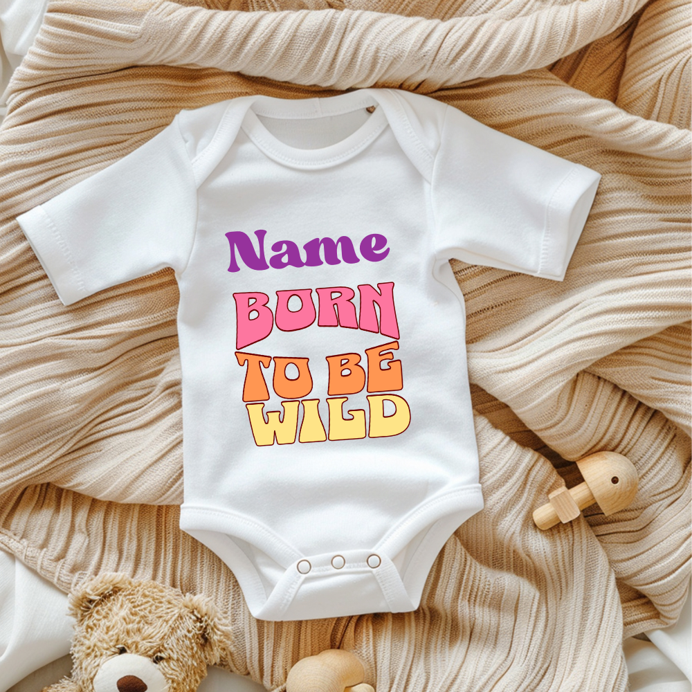 Ein weißer Baby-Body mit dem personalisierbaren Schriftzug 'Name' in lila und darunter den Worten 'BORN TO BE WILD' in einem Retro-Stil in Pink, Orange und Gelb. Der Body liegt auf einer beigen, gestrickten Decke, umgeben von Spielzeug, was eine gemütliche Atmosphäre schafft. Ein auffälliges und personalisierbares Geschenk für kleine, wilde Abenteurer.