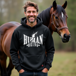 Ein Mann mit einem schwarzen Hoodie, auf dem das Design "Human Being" in großen weißen Buchstaben und mit einem Blitz-Motiv abgebildet ist. Im Hintergrund steht ein braunes Pferd auf einer grünen Wiese.