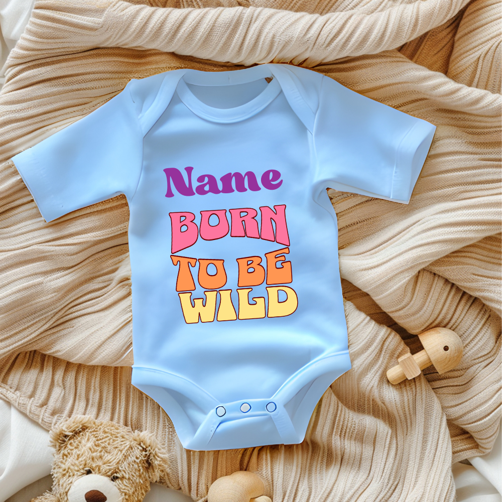 Ein hellblauer Baby-Body mit dem personalisierbaren Schriftzug 'Name' in lila und darunter den Worten 'BORN TO BE WILD' in einem Retro-Stil in Pink, Orange und Gelb. Der Body liegt auf einer beigen, gestrickten Decke, umgeben von Spielzeug, was eine gemütliche Atmosphäre schafft. Ein auffälliges und personalisierbares Geschenk für kleine, wilde Abenteurer.