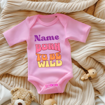 Ein rosa Baby-Body mit dem personalisierbaren Schriftzug 'Name' in lila und darunter den Worten 'BORN TO BE WILD' in einem Retro-Stil in Pink, Orange und Gelb. Der Body liegt auf einer beigen, gestrickten Decke, umgeben von Spielzeug, was eine gemütliche Atmosphäre schafft. Ein auffälliges und personalisierbares Geschenk für kleine, wilde Abenteurer.