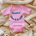 Ein rosa Baby-Body mit dem personalisierbaren Text 'Name' in großer, grüner Schrift und dem darunter geschwungenen Schriftzug 'Born to be Wild' in verspielter, dunkler Schrift. Der Body liegt auf einer beigen, gestrickten Decke und wird von Spielzeug umrahmt, was eine gemütliche Atmosphäre schafft. Ein fröhliches und individuelles Geschenk für kleine Abenteurer.