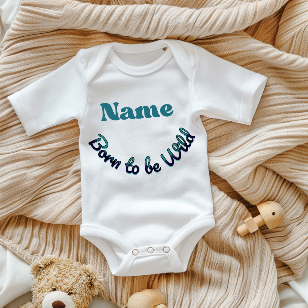 Ein weißer Baby-Body mit dem personalisierbaren Text 'Name' in großer, grüner Schrift und dem darunter geschwungenen Schriftzug 'Born to be Wild' in verspielter, dunkler Schrift. Der Body liegt auf einer beigen, gestrickten Decke und wird von Spielzeug umrahmt, was eine gemütliche Atmosphäre schafft. Ein fröhliches und individuelles Geschenk für kleine Abenteurer.