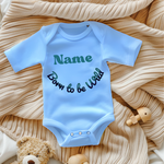 Ein hellblauer Baby-Body mit dem personalisierbaren Text 'Name' in großer, grüner Schrift und dem darunter geschwungenen Schriftzug 'Born to be Wild' in verspielter, dunkler Schrift. Der Body liegt auf einer beigen, gestrickten Decke und wird von Spielzeug umrahmt, was eine gemütliche Atmosphäre schafft. Ein fröhliches und individuelles Geschenk für kleine Abenteurer.