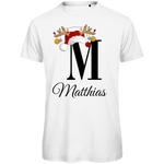 Ein weißes T-Shirt mit einem weihnachtlichen Design. Der große Buchstabe "M" ist mittig auf der Brust abgebildet und trägt einen Weihnachtsmann-Hut sowie Rentiergeweihe, an denen bunte Weihnachtskugeln hängen. Unterhalb des Buchstabens steht der Name "Matthias" in eleganter, geschwungener Schrift. Das Design vermittelt eine festliche Stimmung und ist personalisiert für den Namen Matthias.