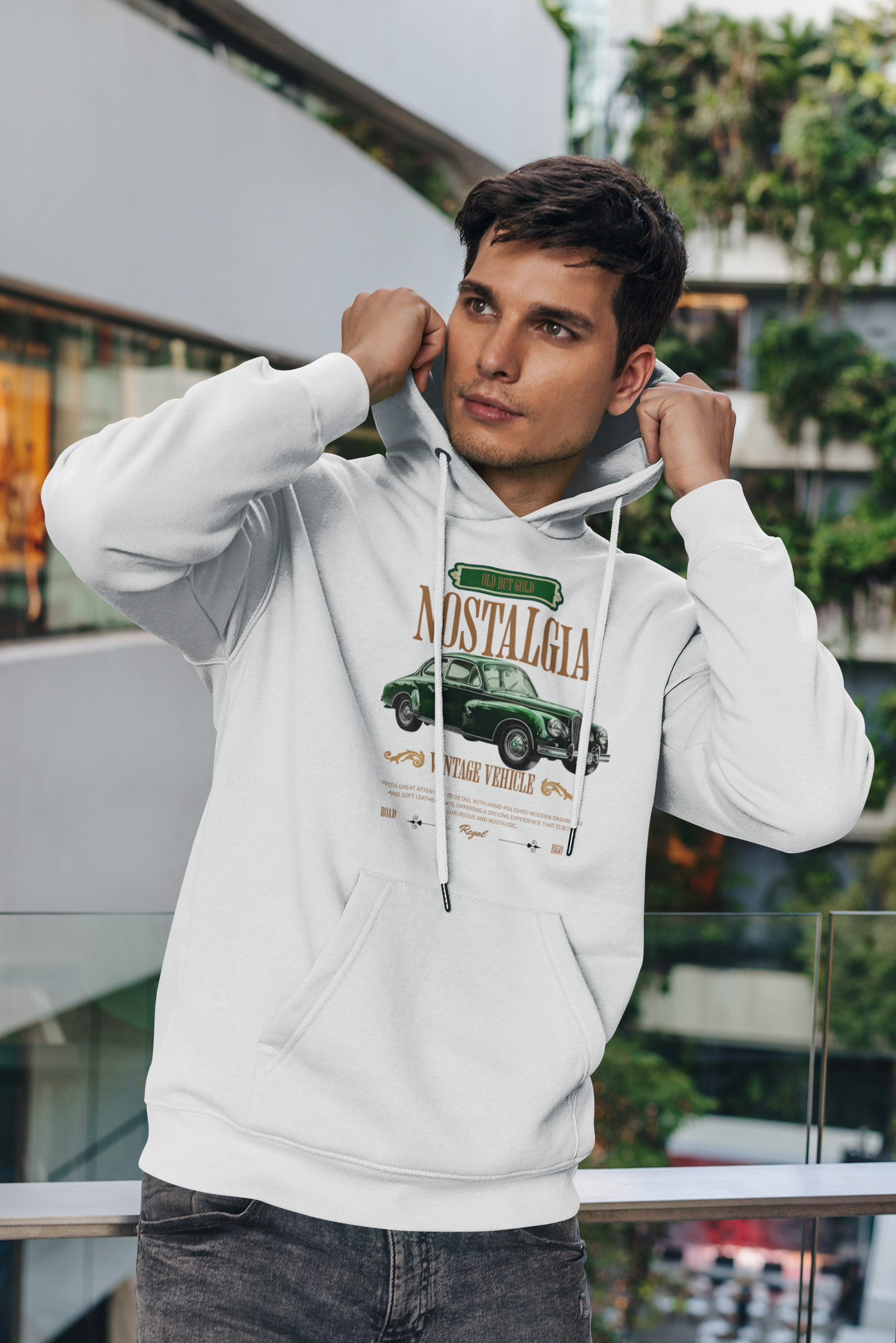 Ein Mann im weißen Hoodie mit einem nostalgischen Vintage-Design. Das Motiv zeigt ein klassisches grünes Auto und den Schriftzug "Nostalgia" darüber. Darunter steht "Vintage Vehicle" mit weiteren dekorativen Elementen und kleinen Texten, die den Retro-Look verstärken. Der Mann hält die Kapuze, während er in die Ferne schaut, was eine entspannte und reflektierende Stimmung vermittelt.