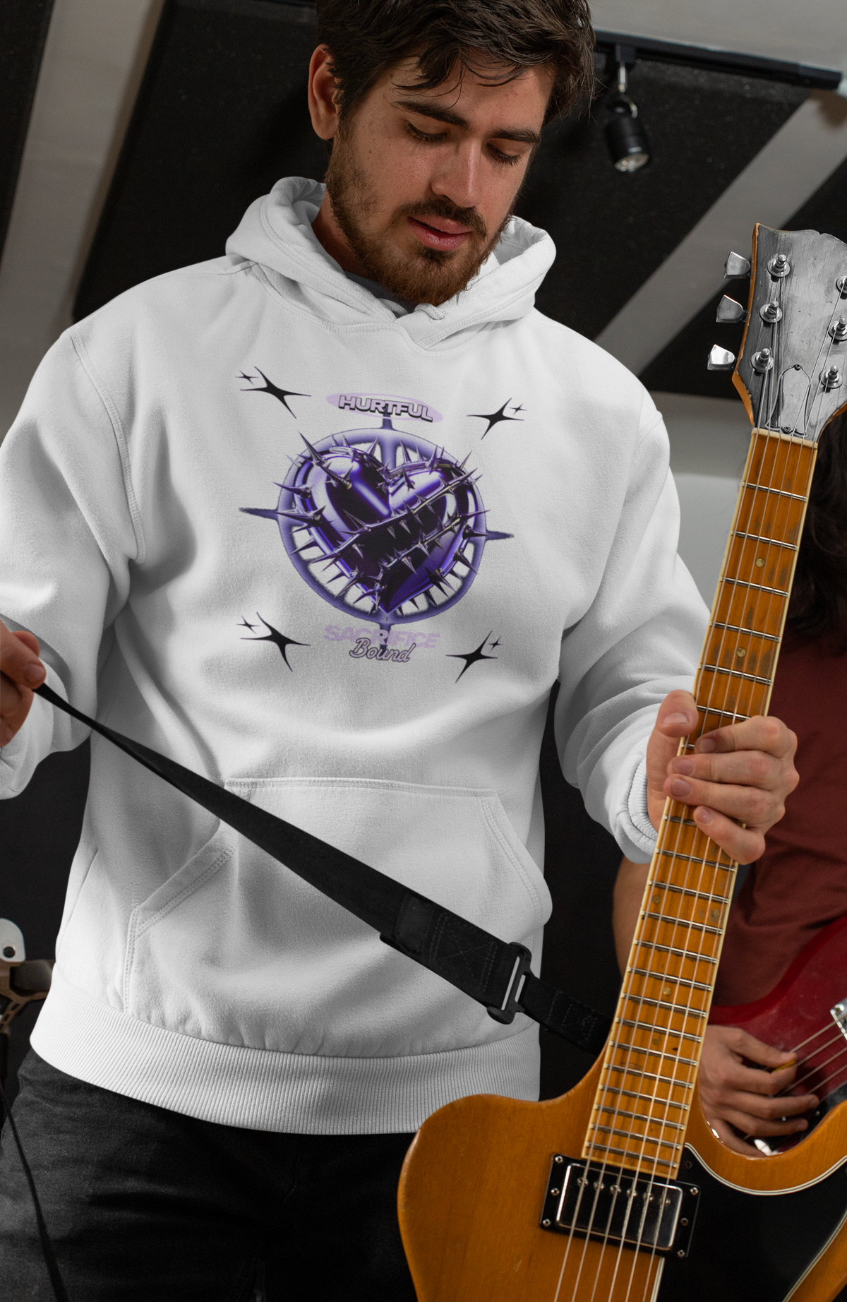 Ein Mann im weißen Hoodie mit einem auffälligen, violetten Herz-Design, das von Dornen umgeben ist. Über dem Herz steht das Wort "Hurtful", darunter "Sacrifice Bound". Der Mann hält eine Gitarre und bereitet sich auf das Spielen vor. Der Hintergrund deutet auf ein Musikstudio hin, was eine rockige und emotionale Atmosphäre vermittelt.