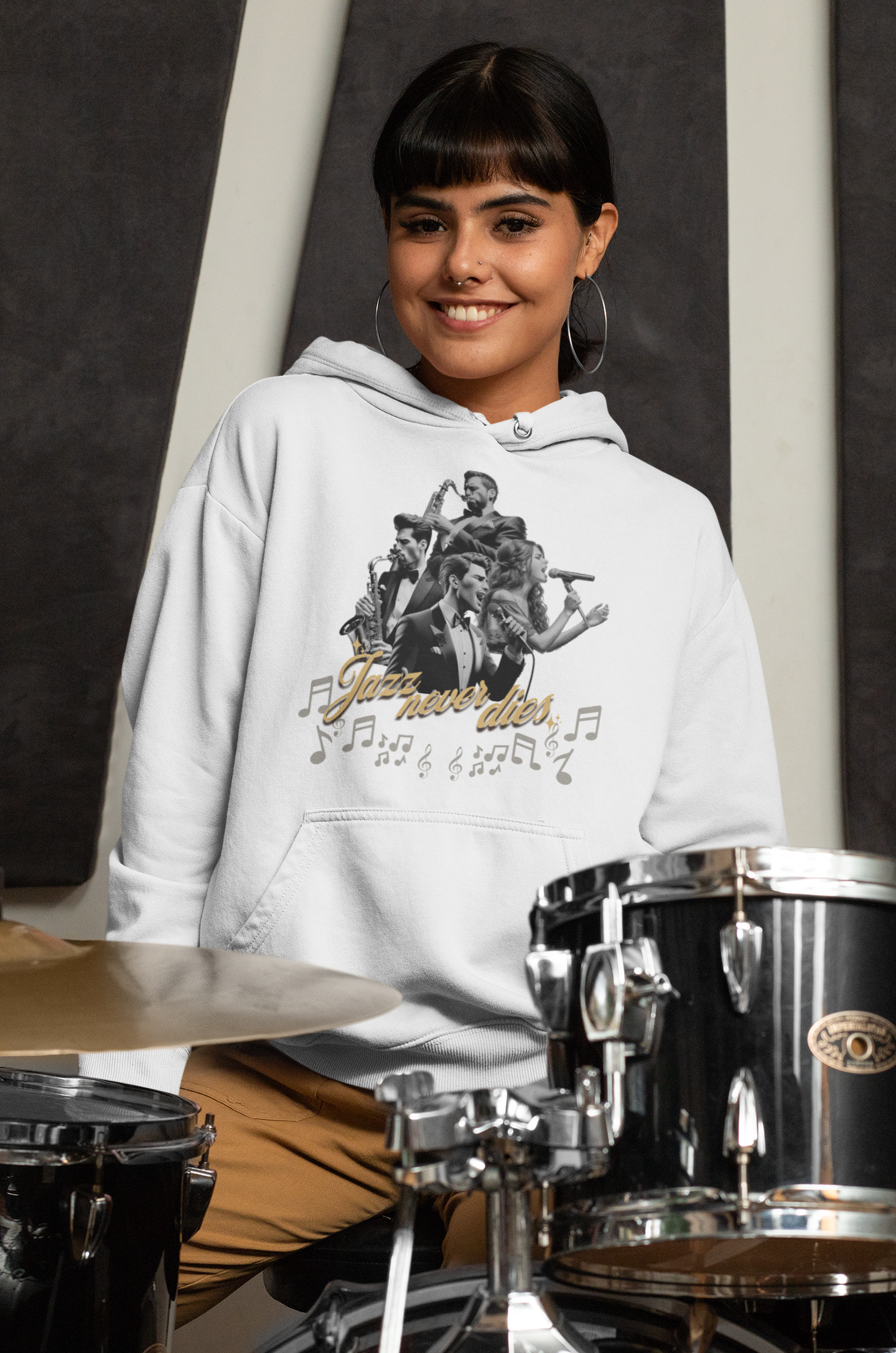 Eine lächelnde Frau, die einen weißen Hoodie trägt, auf dem ein stilvolles Jazz-Design zu sehen ist. Das Design zeigt eine Gruppe von Musikern mit verschiedenen Instrumenten, umgeben von Musiknoten und dem Schriftzug "Jazz never dies" in goldener Schrift. Im Hintergrund sind Schlagzeuge zu sehen, was eine musikalische Atmosphäre vermittelt.