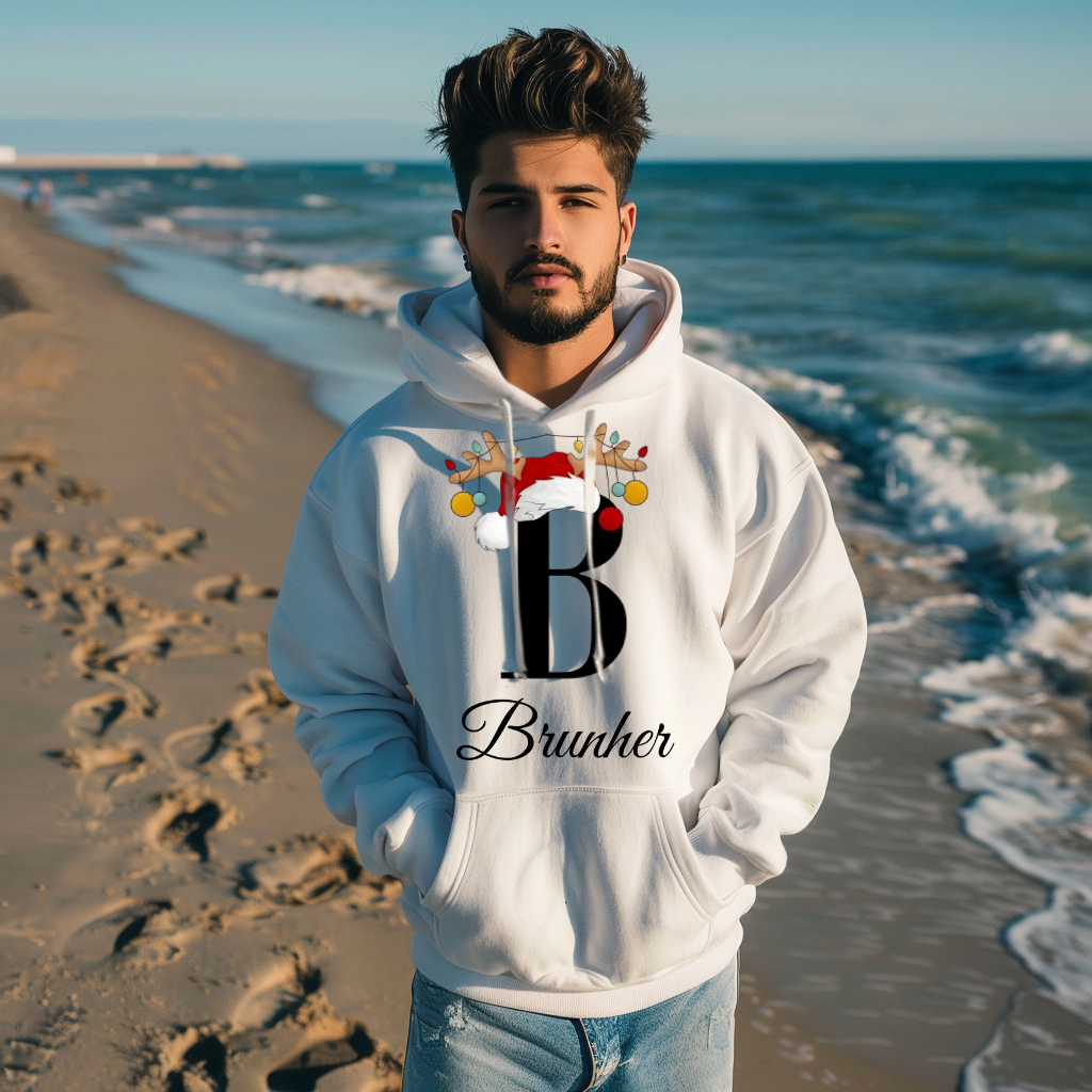 Ein junger Mann steht am Strand und trägt einen weißen Hoodie mit einem weihnachtlichen Design. Der Buchstabe "B" in schwarz ist mit einem Weihnachtsmann-Hut und Rentiergeweihen dekoriert, an denen bunte Kugeln hängen. Unter dem Buchstaben ist der personalisierte Name "Brunher" in eleganter, geschwungener Schrift zu sehen. Der Hoodie strahlt eine festliche Stimmung aus und ist perfekt für die Winterzeit.