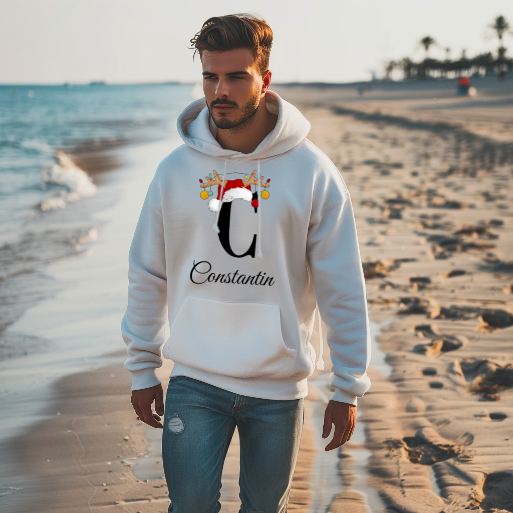 Ein junger Mann spaziert am Strand entlang und trägt einen weißen Hoodie mit einem weihnachtlichen Design. Auf dem Hoodie ist der Buchstabe "C" zu sehen, verziert mit einem Weihnachtsmann-Hut und Rentiergeweihen, an denen bunte Kugeln hängen. Darunter steht der personalisierte Name "Constantin" in einer eleganten, geschwungenen Schrift. Die Szene kombiniert eine entspannte Strandatmosphäre mit einem festlichen, personalisierten Kleidungsstück.