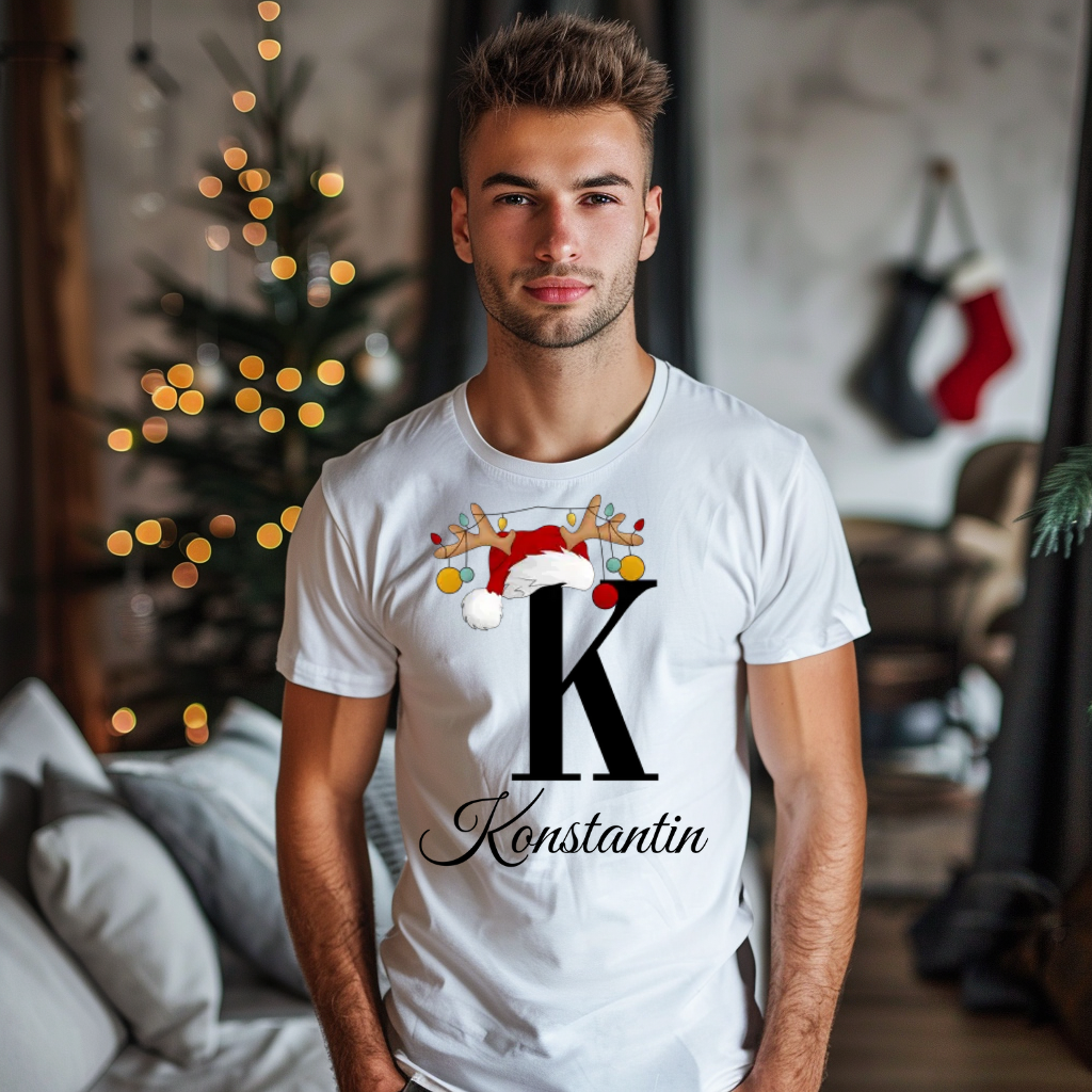 Ein junger Mann steht in einem weihnachtlich dekorierten Raum, im Hintergrund ist ein Weihnachtsbaum mit Lichtern zu sehen. Er trägt ein weißes T-Shirt mit einem festlichen Design. Auf dem T-Shirt ist ein großer Buchstabe "K" abgebildet, der mit einem Weihnachtsmann-Hut und Rentiergeweihen dekoriert ist, an denen bunte Kugeln hängen. Unter dem Buchstaben steht der Name "Konstantin" in eleganter, geschwungener Schrift. Das Design strahlt eine personalisierte und festliche Stimmung aus.