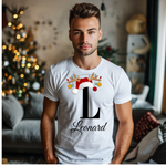 Ein junger Mann steht in einem weihnachtlich dekorierten Raum, im Hintergrund ist ein Weihnachtsbaum mit Lichtern zu sehen. Er trägt ein weißes T-Shirt mit einem festlichen Design. Auf dem T-Shirt ist ein großer Buchstabe "L" abgebildet, der mit einem Weihnachtsmann-Hut und Rentiergeweihen dekoriert ist, an denen bunte Kugeln hängen. Unter dem Buchstaben steht der Name "Leonard" in eleganter, geschwungener Schrift. Das Design vermittelt eine personalisierte und festliche Stimmung.