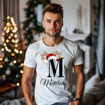 Ein junger Mann steht in einem weihnachtlich dekorierten Raum, im Hintergrund ist ein geschmückter Weihnachtsbaum zu sehen. Er trägt ein weißes T-Shirt mit einem festlichen Design. Auf dem T-Shirt ist ein großer Buchstabe "M" abgebildet, der mit einem Weihnachtsmann-Hut und Rentiergeweihen dekoriert ist, an denen bunte Kugeln hängen. Unter dem Buchstaben steht der Name "Matthias" in eleganter, geschwungener Schrift. Das Design vermittelt eine personalisierte und festliche Stimmung.