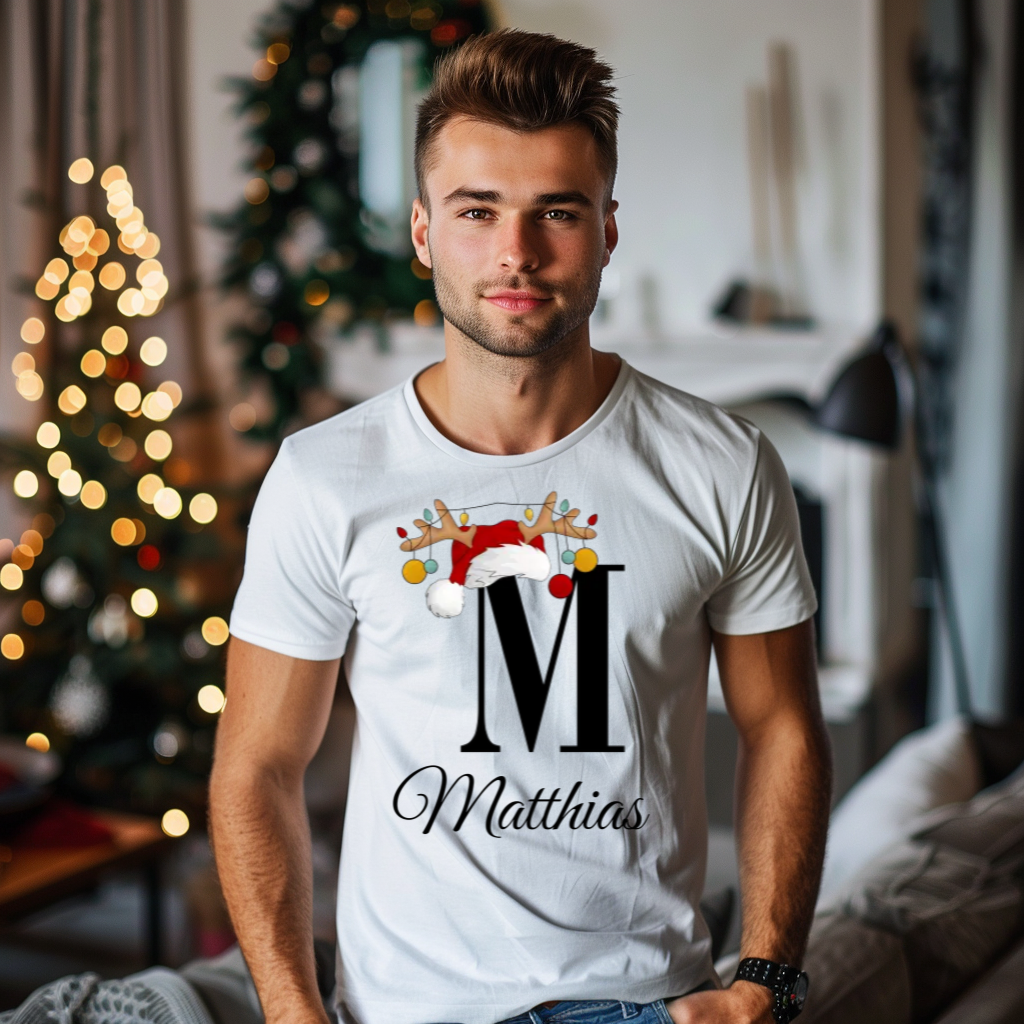 Ein junger Mann steht in einem weihnachtlich dekorierten Raum, im Hintergrund ist ein geschmückter Weihnachtsbaum zu sehen. Er trägt ein weißes T-Shirt mit einem festlichen Design. Auf dem T-Shirt ist ein großer Buchstabe "M" abgebildet, der mit einem Weihnachtsmann-Hut und Rentiergeweihen dekoriert ist, an denen bunte Kugeln hängen. Unter dem Buchstaben steht der Name "Matthias" in eleganter, geschwungener Schrift. Das Design vermittelt eine personalisierte und festliche Stimmung.