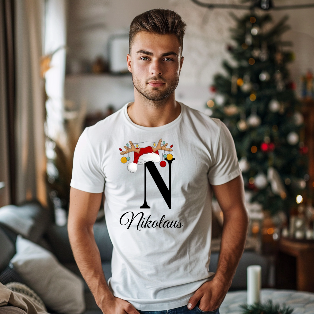 Ein junger Mann steht in einem weihnachtlich dekorierten Raum, im Hintergrund ist ein geschmückter Weihnachtsbaum zu sehen. Er trägt ein weißes T-Shirt mit einem festlichen Design. Auf dem T-Shirt ist ein großer Buchstabe "N" abgebildet, der mit einem Weihnachtsmann-Hut und Rentiergeweihen dekoriert ist, an denen bunte Kugeln hängen. Unter dem Buchstaben steht der Name "Nikolaus" in eleganter, geschwungener Schrift. Das Design vermittelt eine personalisierte und festliche Stimmung.