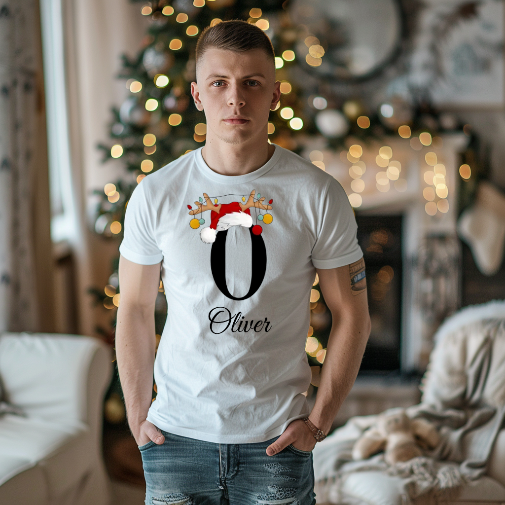 Ein junger Mann steht in einem weihnachtlich dekorierten Raum, im Hintergrund ist ein geschmückter Weihnachtsbaum und ein Kamin zu sehen. Er trägt ein weißes T-Shirt mit einem festlichen Design. Auf dem T-Shirt ist ein großer Buchstabe "O" abgebildet, der mit einem Weihnachtsmann-Hut und Rentiergeweihen dekoriert ist, an denen bunte Kugeln hängen. Unter dem Buchstaben steht der Name "Oliver" in eleganter, geschwungener Schrift. Das Design strahlt eine personalisierte und weihnachtliche Stimmung aus.