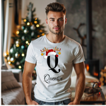 Ein junger Mann steht in einem weihnachtlich dekorierten Raum, im Hintergrund ist ein geschmückter Weihnachtsbaum und ein Kamin zu sehen. Er trägt ein weißes T-Shirt mit einem festlichen Design. Auf dem T-Shirt ist ein großer Buchstabe "Q" abgebildet, der mit einem Weihnachtsmann-Hut und Rentiergeweihen dekoriert ist, an denen bunte Kugeln hängen. Unter dem Buchstaben steht der Name "Quentin" in eleganter, geschwungener Schrift. Das Design vermittelt eine personalisierte und festliche Stimmung.