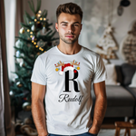 Ein junger Mann steht in einem weihnachtlich dekorierten Raum, im Hintergrund ist ein geschmückter Weihnachtsbaum zu sehen. Er trägt ein weißes T-Shirt mit einem festlichen Design. Auf dem T-Shirt ist ein großer Buchstabe "R" abgebildet, der mit einem Weihnachtsmann-Hut und Rentiergeweihen dekoriert ist, an denen bunte Kugeln hängen. Unter dem Buchstaben steht der Name "Rudolf" in eleganter, geschwungener Schrift. Das Design vermittelt eine personalisierte und festliche Stimmung.