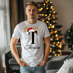 Ein junger Mann steht in einem weihnachtlich dekorierten Raum, im Hintergrund ist ein geschmückter Weihnachtsbaum zu sehen. Er trägt ein weißes T-Shirt mit einem festlichen Design. Auf dem T-Shirt ist ein großer Buchstabe "T" abgebildet, der mit einem Weihnachtsmann-Hut und Rentiergeweihen dekoriert ist, an denen bunte Kugeln hängen. Unter dem Buchstaben steht der Name "Thorsten" in eleganter, geschwungener Schrift. Das Design vermittelt eine personalisierte und festliche Stimmung.