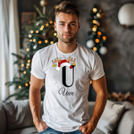 Ein junger Mann steht in einem weihnachtlich dekorierten Raum, im Hintergrund ist ein geschmückter Weihnachtsbaum zu sehen. Er trägt ein weißes T-Shirt mit einem festlichen Design. Auf dem T-Shirt ist ein großer Buchstabe "U" abgebildet, der mit einem Weihnachtsmann-Hut und Rentiergeweihen dekoriert ist, an denen bunte Kugeln hängen. Unter dem Buchstaben steht der Name "Uwe" in eleganter, geschwungener Schrift. Das Design vermittelt eine personalisierte und festliche Stimmung.