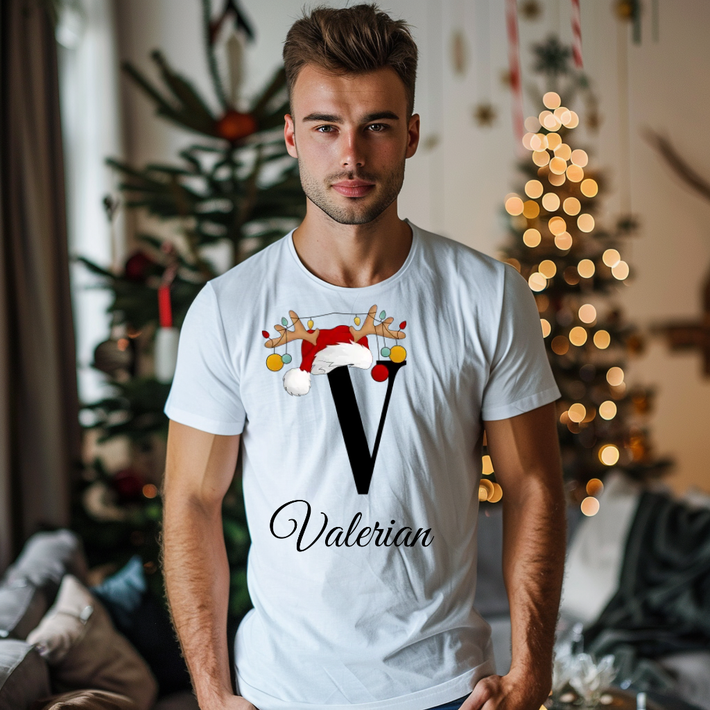 Ein junger Mann steht in einem weihnachtlich dekorierten Raum, im Hintergrund ist ein geschmückter Weihnachtsbaum mit Lichtern zu sehen. Er trägt ein weißes T-Shirt mit einem festlichen Design. Auf dem T-Shirt ist ein großer Buchstabe "V" abgebildet, der mit einem Weihnachtsmann-Hut und Rentiergeweihen dekoriert ist, an denen bunte Kugeln hängen. Unter dem Buchstaben steht der Name "Valerian" in eleganter, geschwungener Schrift. Das Design strahlt eine personalisierte und festliche Stimmung aus.