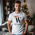 Ein junger Mann steht in einem gemütlichen, weihnachtlich dekorierten Raum, im Hintergrund ist ein geschmückter Weihnachtsbaum zu sehen. Er trägt ein weißes T-Shirt mit einem festlichen Design. Auf dem T-Shirt ist ein großer Buchstabe "W" abgebildet, der mit einem Weihnachtsmann-Hut und Rentiergeweihen dekoriert ist, an denen bunte Kugeln hängen. Unter dem Buchstaben steht der Name "William" in eleganter, geschwungener Schrift. Das Design strahlt eine personalisierte und festliche Stimmung aus.