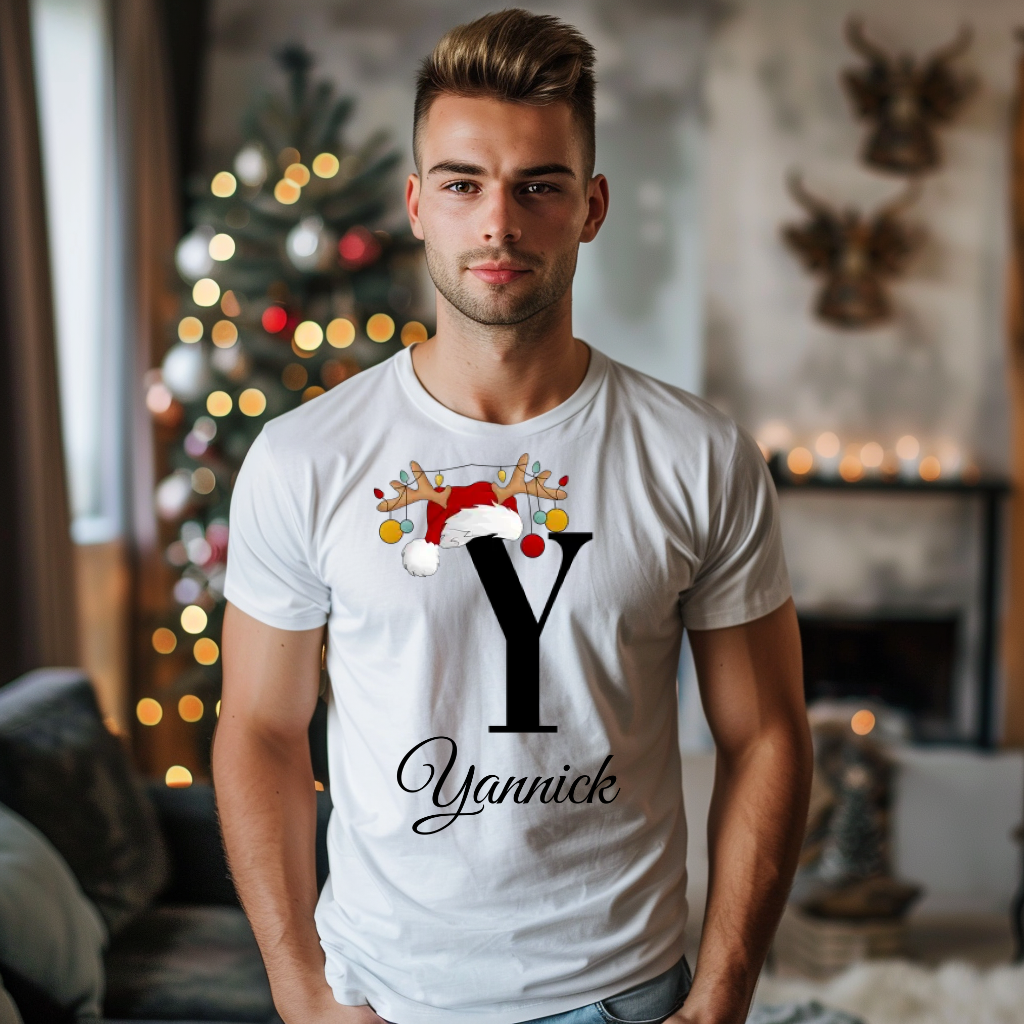 Ein weißes T-Shirt mit einem festlichen Design, das den Buchstaben „Y“ zeigt. Der Buchstabe ist schwarz und wird mit einem Weihnachtsmann-Hut und Rentiergeweihen geschmückt, an denen bunte Kugeln hängen. Unter dem Buchstaben steht der Name „Yannick“ in eleganter, geschwungener Schrift. Im Hintergrund ist ein weihnachtlich dekorierter Raum zu sehen, was die festliche Stimmung verstärkt.