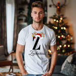 Ein weißes T-Shirt mit einem weihnachtlichen Design, das den Buchstaben „Z“ in schwarzer Farbe zeigt. Der Buchstabe ist mit einem Weihnachtsmann-Hut und Rentiergeweihen verziert, an denen bunte Kugeln hängen. Unter dem Buchstaben steht der Name „Zacharias“ in eleganter, geschwungener Schrift. Der Mann trägt das T-Shirt in einem gemütlichen Wohnzimmer mit einem festlich dekorierten Weihnachtsbaum im Hintergrund, was die weihnachtliche Stimmung unterstreicht.