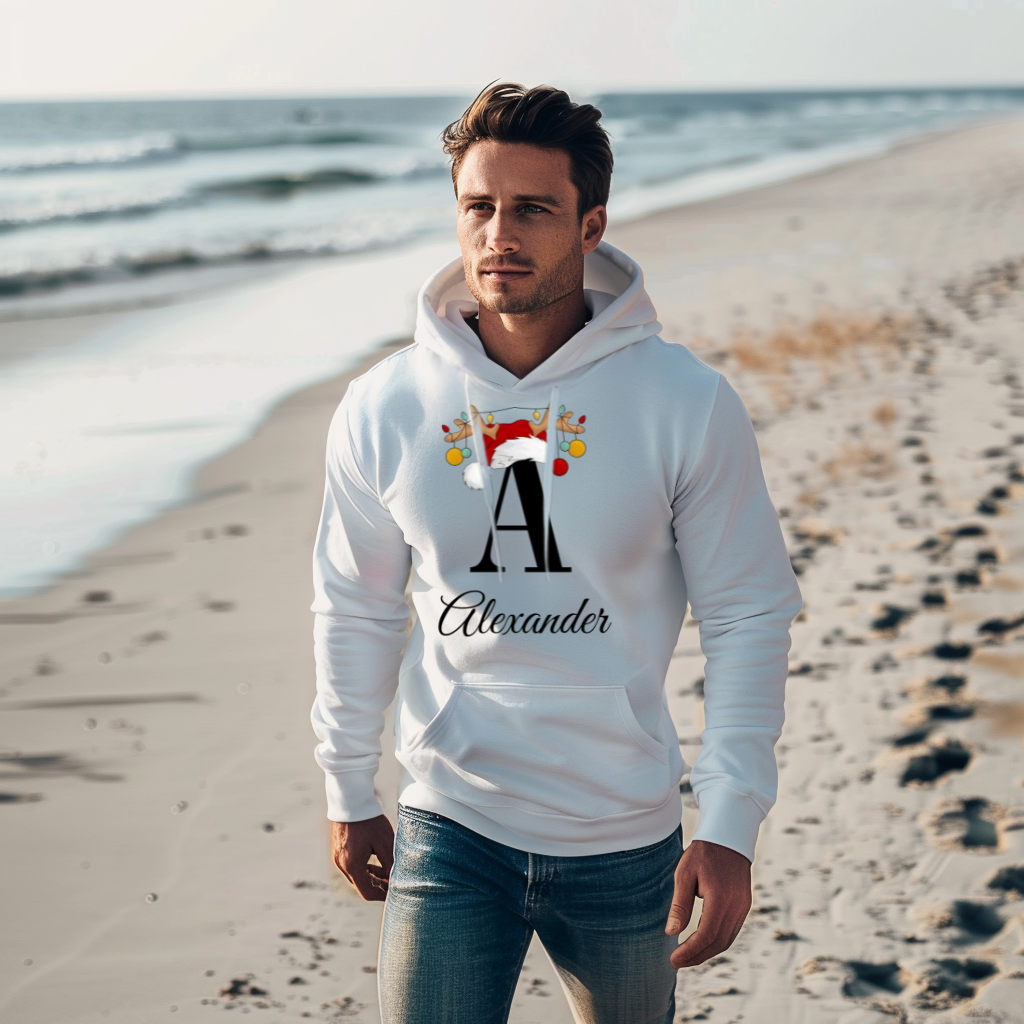 Ein Mann steht an einem Strand und trägt einen weißen Hoodie mit einem weihnachtlichen Design. Auf dem Hoodie ist ein großes, schwarzes "A" zu sehen, das mit einem Weihnachtsmann-Hut und Rentiergeweihen dekoriert ist, an denen bunte Kugeln hängen. Unter dem Buchstaben steht der Name "Alexander" in eleganter, geschwungener Schrift. Der Hoodie kombiniert einen festlichen Stil mit einer personalisierten Note.