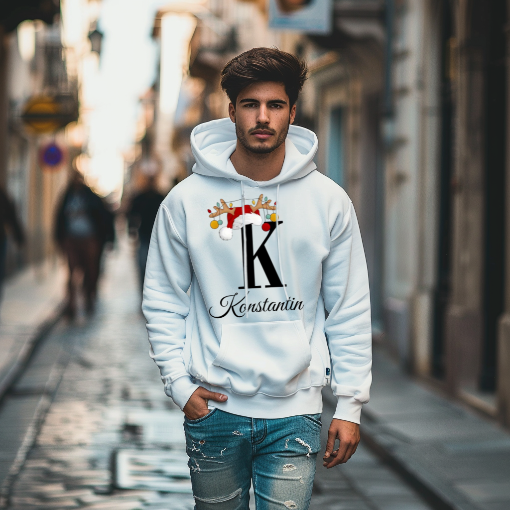 Ein junger Mann trägt einen weißen Hoodie mit einem weihnachtlichen Design. Der Buchstabe "K" ist prominent in Schwarz abgebildet und wird mit einem Weihnachtsmann-Hut sowie Rentiergeweihen geschmückt, an denen bunte Kugeln hängen. Darunter ist der personalisierte Name "Konstantin" in eleganter, geschwungener Schrift zu sehen. Die Szenerie zeigt eine winterliche, festliche Stimmung und der Hoodie vermittelt eine perfekt personalisierte Note für die Weihnachtszeit.