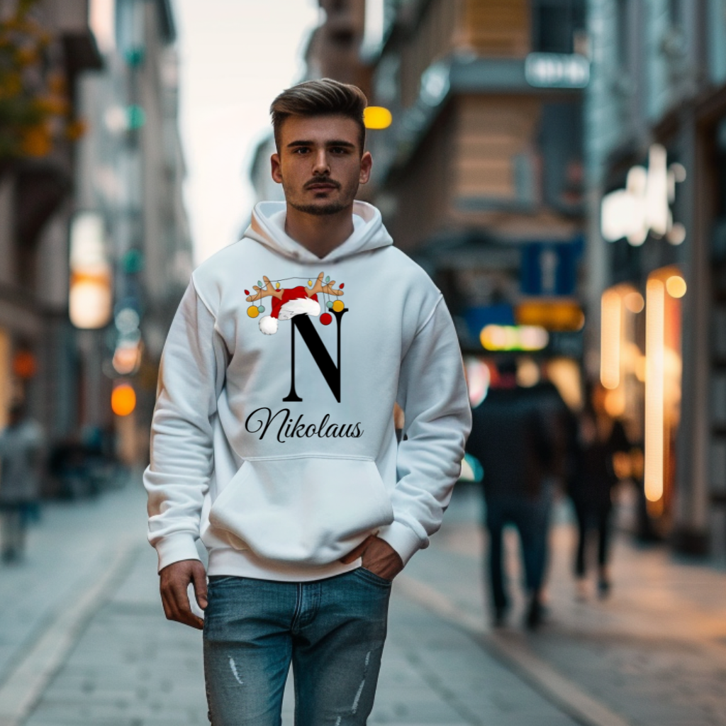 Ein weißer Hoodie mit einem weihnachtlichen Design. Der Buchstabe "N" wird durch eine Weihnachtsmütze und Rentiergeweihe verziert, an denen bunte Christbaumkugeln hängen. Unter dem Buchstaben steht der Name "Nikolaus" in eleganter, geschwungener Schrift. Perfekt für die festliche Jahreszeit und personalisiert für eine besondere Note.