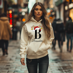 Auf dem Bild ist eine junge Frau zu sehen, die einen cremefarbenen Hoodie trägt. Auf dem Hoodie ist ein festliches Design mit einem großen schwarzen Buchstaben „B“ sowie dem Namen „Bernadette“ in eleganter Schrift gedruckt. Über dem Buchstaben sind ein Rentiergeweih, ein Weihnachtsmann-Hut und bunte Kugeln als weihnachtliche Dekoration angebracht. Die Frau steht in einer belebten, unscharf abgebildeten Stadtstraße mit Lichtern und Menschen im Hintergrund, die eine winterliche, stimmungsvolle Atmosphäre verm