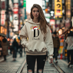 Das Bild zeigt eine junge Frau in einem weißen Hoodie mit einem festlichen, personalisierten Design. Im Zentrum des Designs steht der Buchstabe „D“, verziert mit einem Rentiergeweih, einem Weihnachtsmann-Hut und bunten Kugeln, die eine weihnachtliche Atmosphäre erzeugen. Unter dem Buchstaben ist der Name „Danielle“ in einer eleganten, geschwungenen Schrift abgebildet. Die Szenerie wirkt urban und lebendig, passend zur stilvollen und individuellen Gestaltung des Hoodies für die Winter- und Weihnachtszeit.