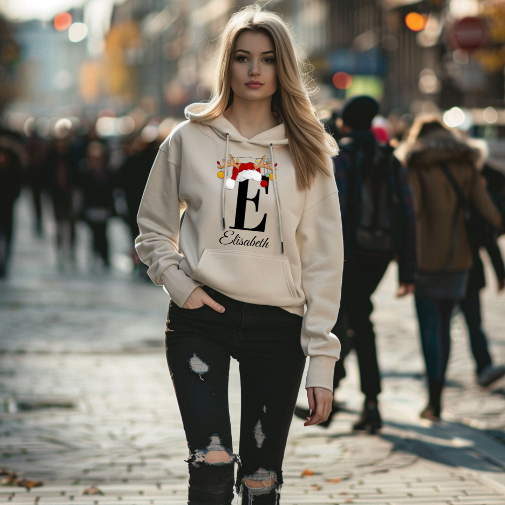 Auf dem Bild ist eine junge Frau zu sehen, die einen weißen Hoodie trägt. Auf dem Hoodie ist ein festliches Design mit einem großen schwarzen Buchstaben sowie einem Namen passend zum Buchstaben, in eleganter Schrift gedruckt. Über dem Buchstaben sind ein Rentiergeweih, ein Weihnachtsmann-Hut und bunte Kugeln als weihnachtliche Dekoration angebracht. Die Frau steht in einer belebten, unscharf abgebildeten Stadtstraße mit Lichtern und Menschen im Hintergrund, die eine winterliche, stimmungsvolle Atmosphäre ve