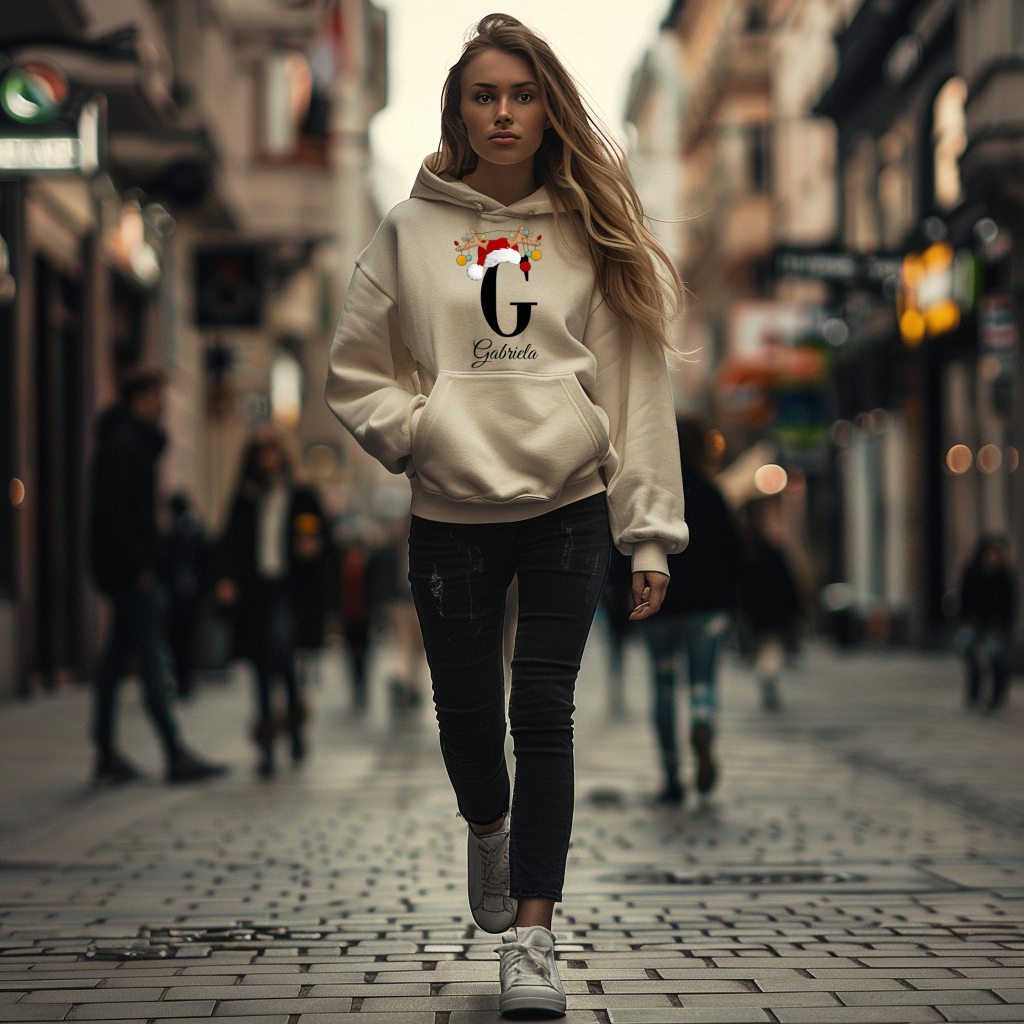 Auf dem Bild ist eine junge Frau zu sehen, die einen weißen Hoodie trägt. Auf dem Hoodie ist ein festliches Design mit einem großen schwarzen Buchstaben sowie der Namen „Gabriela“ passend zum Buchstaben, in eleganter Schrift gedruckt. Über dem Buchstaben sind ein Rentiergeweih, ein Weihnachtsmann-Hut und bunte Kugeln als weihnachtliche Dekoration angebracht. Die Frau steht in einer belebten, unscharf abgebildeten Stadtstraße mit Lichtern und Menschen im Hintergrund, mit einer winterlichen, Atmosphäre. 