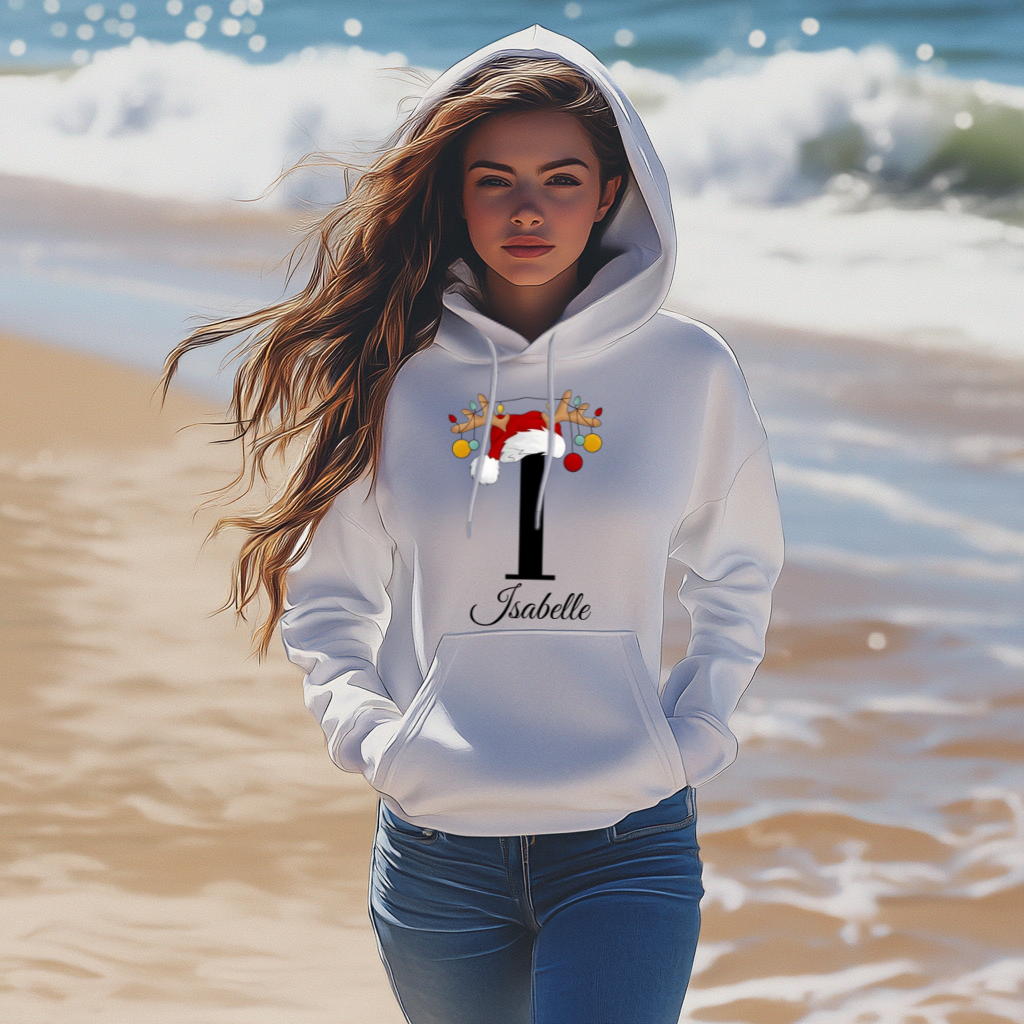 Eine Frau steht am Strand und trägt einen weißen Hoodie mit weihnachtlichem Design. Der Hoodie zeigt den Buchstaben "I", verziert mit einem Weihnachtsmann-Hut und Rentiergeweihen, an denen bunte Kugeln hängen. Unter dem Buchstaben ist der Name "Isabelle" in eleganter, geschwungener Schrift abgebildet. Im Hintergrund sieht man das Meer und sanfte Wellen, die eine entspannte, natürliche Atmosphäre schaffen. Die Frau hat langes, leicht gewelltes Haar, das im Wind weht, und blickt mit einem sanften Ausdruck. 